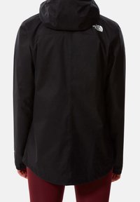 Person, der eine schwarze Kapuzenjacke von North Face trägt, von hinten gezeigt, kombiniert mit dunkelroten Hosen vor einem einfarbigen Hintergrund.