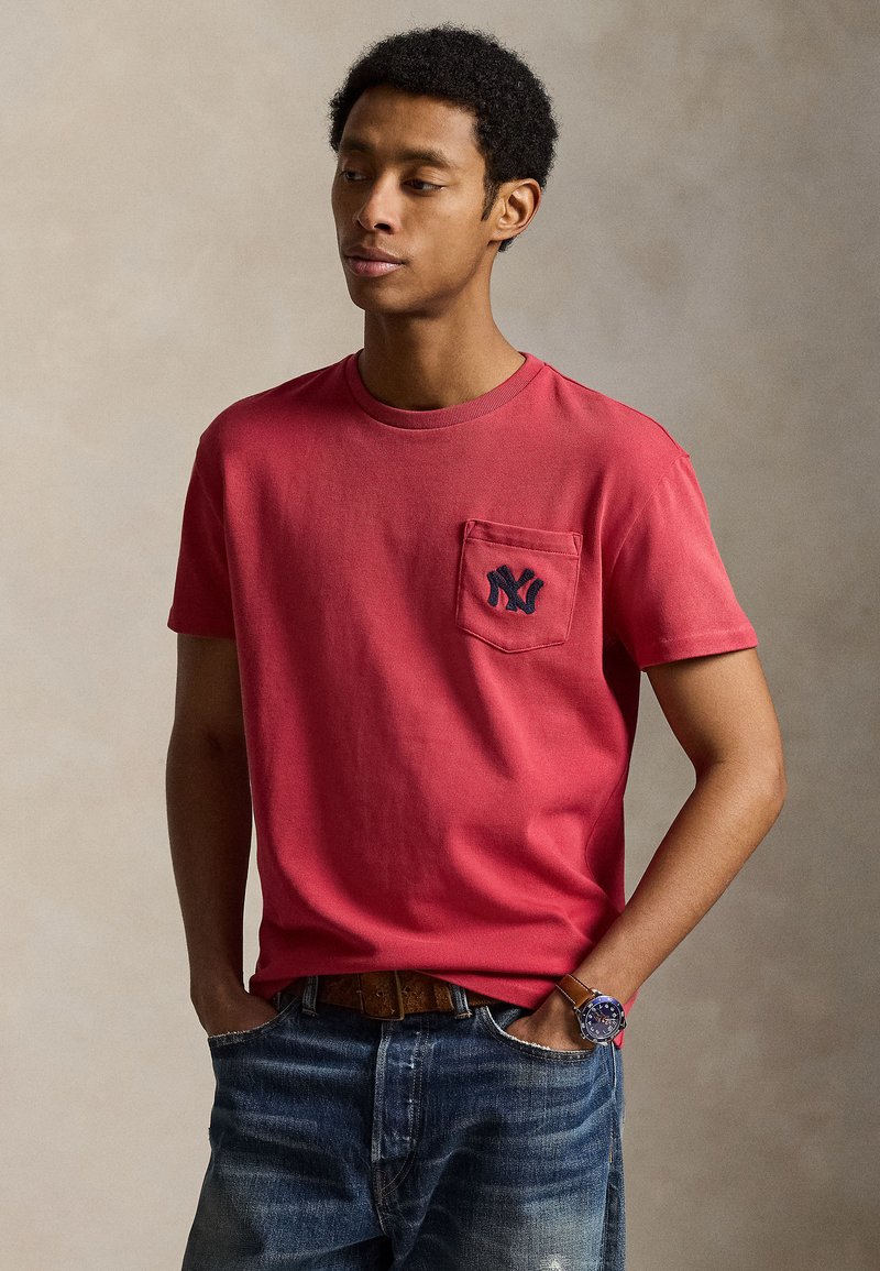 Homme portant un t-shirt rouge avec un logo bleu marine sur la poche, un jean bleu, une ceinture marron et une montre, debout les mains dans les poches.