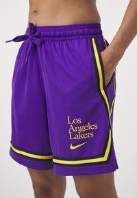 Personne portant un short violet des Los Angeles Lakers avec des bordures jaunes et noires, main gauche dans la poche, logo Nike sous le nom de l'équipe.