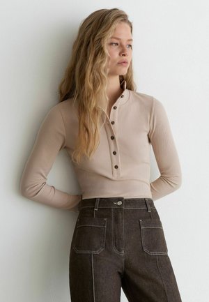 Jonge vrouw met lang blond haar, draagt een beige, knoopjes langemouwen top en hoge taille donkere spijkerbroek, staande tegen een witte muur.