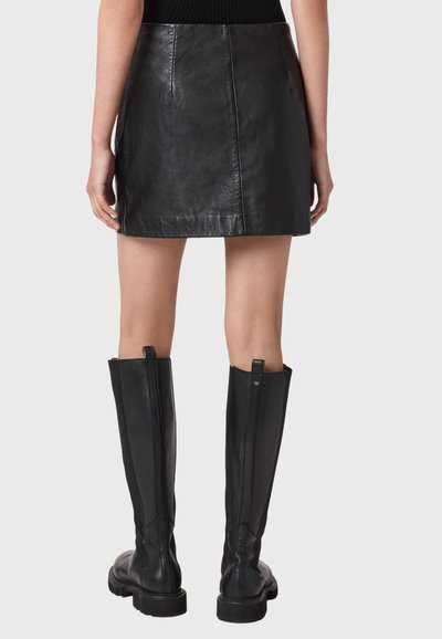 Femme portant une mini-jupe en cuir noire et des bottes en cuir noir montant jusqu'aux genoux, debout, mains détendues le long du corps, vue de dos.