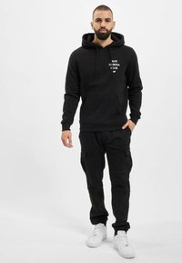 Czarna bluza z kapturem z kieszenią z przodu i białym napisem graficznym, w zestawieniu z czarnymi spodniami cargo i białymi sneakersami. Prosty, casualowy design i dopasowanie.