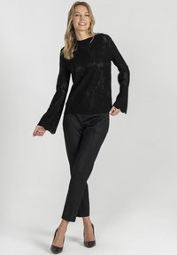 Czarna koronkowa bluzka z długim rękawem i falbaniastym dołem, zestawiona z dopasowanymi czarnymi spodniami i spiczastymi czarnymi szpilkami, na neutralnym tle.