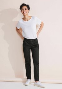 Vit kortärmad skjorta, svarta byxor med framficka, neutral bakgrund, modell i avslappnad pose, bärande vita sneakers.