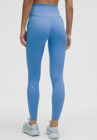 Zilas augsta vidukļa legingi ar gludu tekstūru, sānu kabatām un diskrētu logo pie potītes. Apvienoti ar gaiši pelēkiem sporta apaviem.