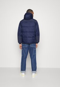 Marinblå puffjacka med huva, med quiltad design, elastiska ärmslut och en något oversized passform, i kombination med ljusblå denimbyxor.