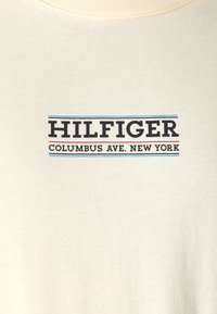 T-shirt color crema con un logo grafico che recita "HILFIGER COLUMBUS AVE. NEW YORK" in testo blu e nero. Tessuto morbido e strutturato.