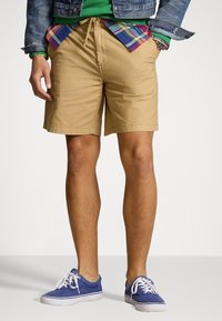 Hombre con pantalones cortos beiges, zapatillas azules, sudadera verde, camisa de cuadros y chaqueta de mezclilla, con las manos en los bolsillos, contra un fondo blanco.