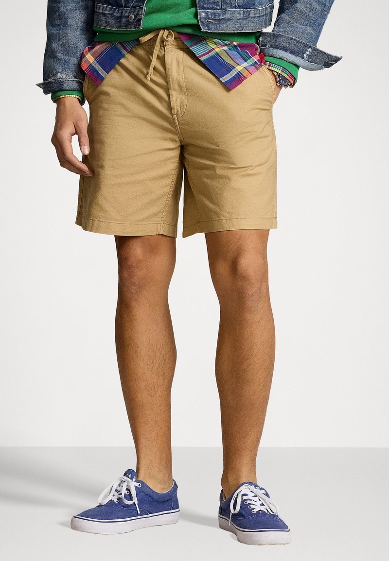 Hombre con pantalones cortos beiges, zapatillas azules, sudadera verde, camisa de cuadros y chaqueta de mezclilla, con las manos en los bolsillos, contra un fondo blanco.