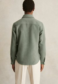 Cămașă din suede verde cu mâneci lungi, manșete cu nasturi și tiv curbat. Textură moale, croială ajustată și design clasic al gulerului.