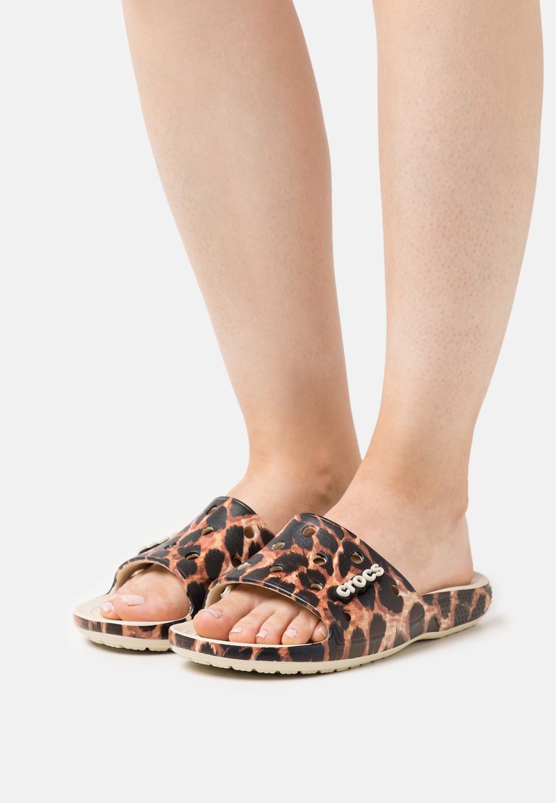 Pieds portant des sandales Crocs à enfiler à imprimé léopard avec les orteils ouverts sur un fond neutre.