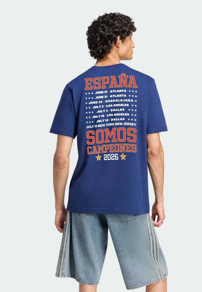 Young man with curly hair wearing a blue T-shirt listing US cities and dates, with text "España Somos Campeones 2026" on the back.
