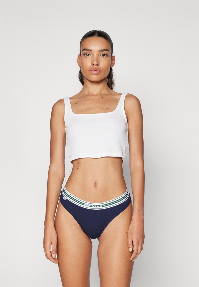 Top court blanc à encolure carrée associé à un slip hipster bleu marine avec une ceinture rayée verte et blanche.