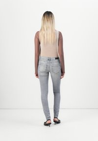 Žena s dlouhými blond vlasy, otočená zády, má na sobě béžový top, hnědé dlouhé rukávy, šedé úzké džíny a černé vysoké podpatky na bílém pozadí.