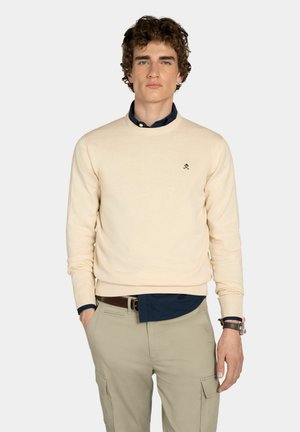 Joven con cabello rizado que lleva un suéter beige de cuello redondo sobre una camisa oscura y pantalones cargo beige, de pie con una mano en el bolsillo.