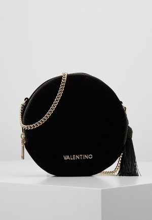 Sac à main rond en velours noir Valentino avec bandoulière chaîne dorée, logo doré, fermeture éclair et gland noir reposant sur une surface blanche.