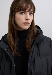 Zwart gewatteerd jack met capuchon, voorzien van een ritssluiting, glanzende afwerking en zijzakken, gedragen over een zwarte coltrui.