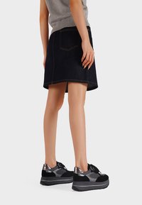 Donna che indossa una gonna in jeans scuri e sneakers platform nere con accenti metallici e grigi, in piedi davanti a uno sfondo semplice.