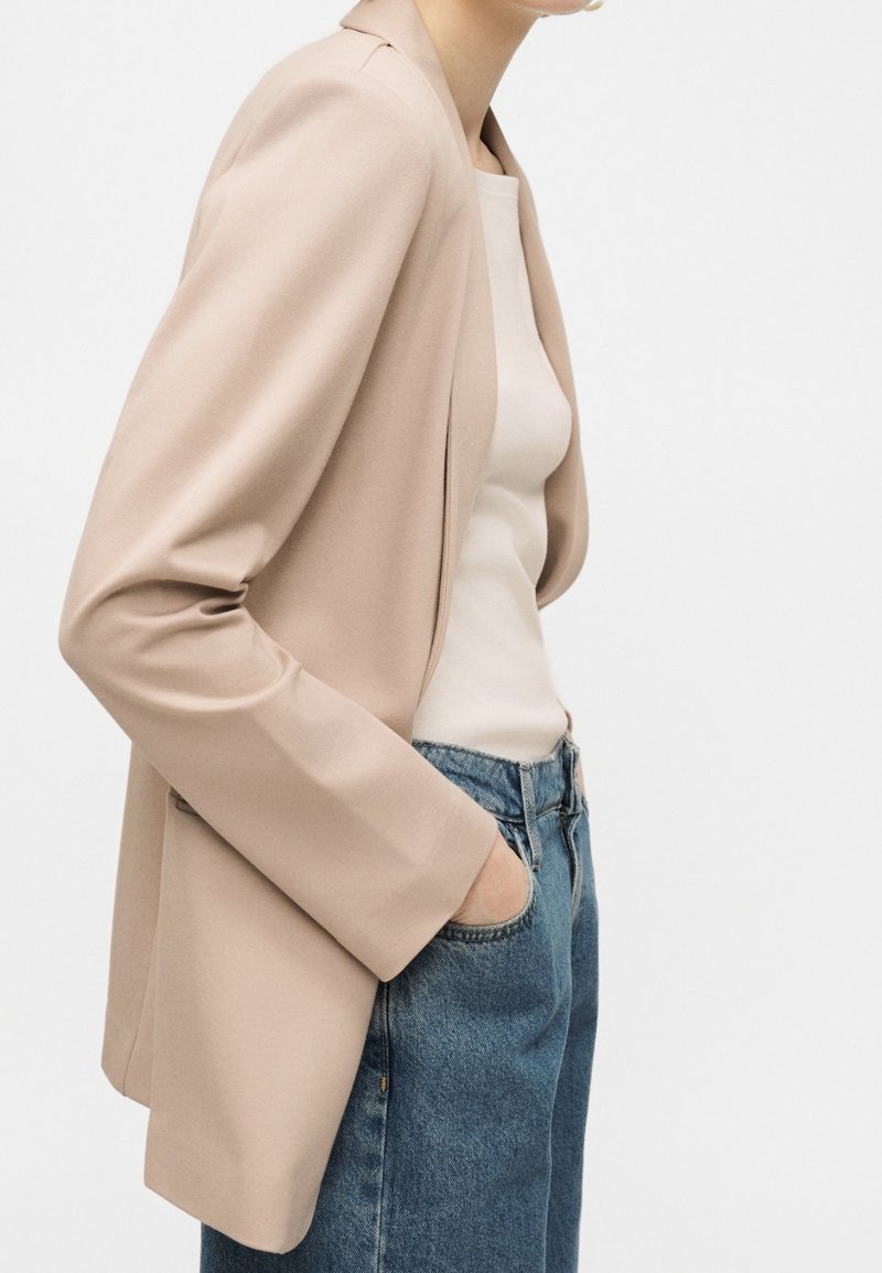 Personne portant un blazer beige sur un haut blanc avec un jean en denim bleu, debout avec une main dans la poche, vue de côté des épaules aux hanches.