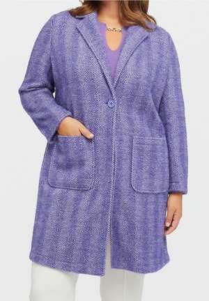 Lang frakke med lilla sildebensmønster med to forlommer, lukning med en enkelt knap, båret over en lilla top og hvide bukser.