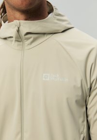 Primo piano di una persona che indossa una giacca con zip e cappuccio Jack Wolfskin di un beige chiaro, con il logo del marchio sul petto.