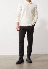 Hombre vistiendo una camisa polo blanca de mangas largas, pantalones ajustados negros y botas de cuero negro con cordones, de pie sobre un suelo de concreto contra una pared blanca.