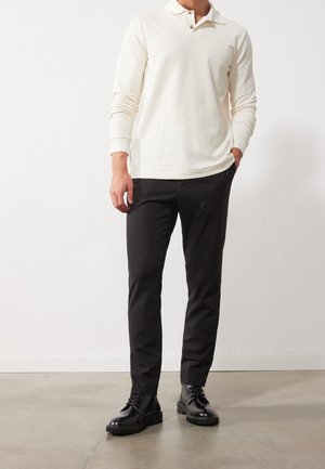 Muž v bílé polo košili s dlouhým rukávem, černých slim-fit kalhotách a černých kožených šněrovacích botách, stojící na betonové podlaze proti bílé zdi.