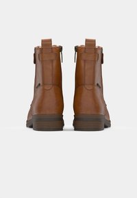 Par bruna läderstövletter med blixtlås på sidan och dragflikar, sedd bakifrån mot en enkel vit bakgrund.