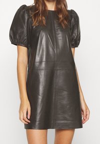 Abito in pelle nera con maniche corte a sbuffo, forma strutturata e design minimalista con cuciture visibili e texture liscia.