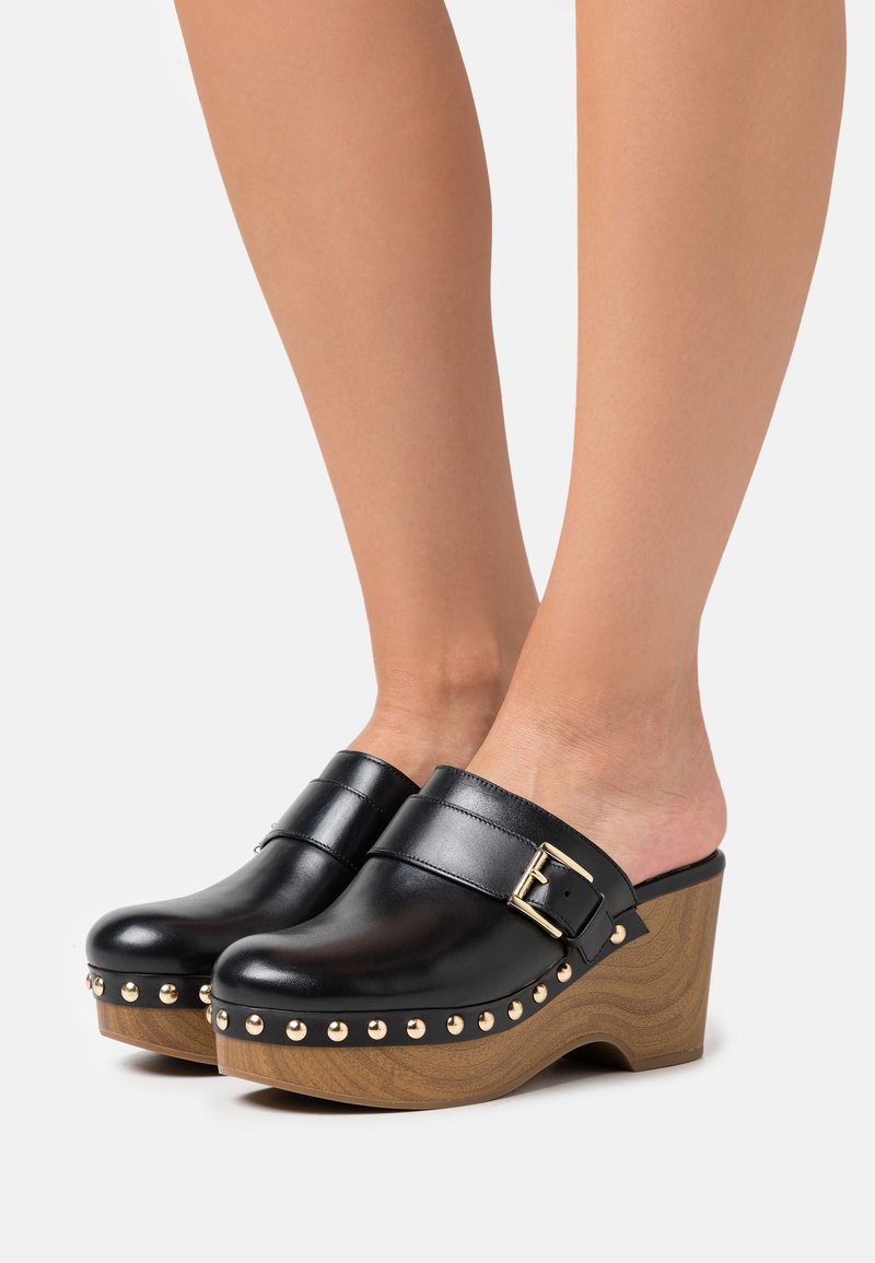 Sabots en cuir noir avec talons compensés en bois, clous dorés le long de la semelle et une bride à boucle dorée portés pieds nus.