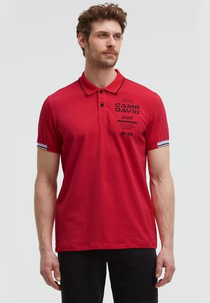 Man wearing a red polo shirt with black text on the chest and black pants, standing against a plain light background.