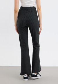 Pantalons évasés noirs et gris à rayures, avec une taille haute, présentant une texture lisse et un subtil éclat, associés à des baskets noires.