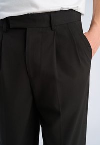 Mand iført sorte plisserede bukser med hånden i lommen og hvid skjorte stoppet ind, beskåret ved talje og midt på låret.