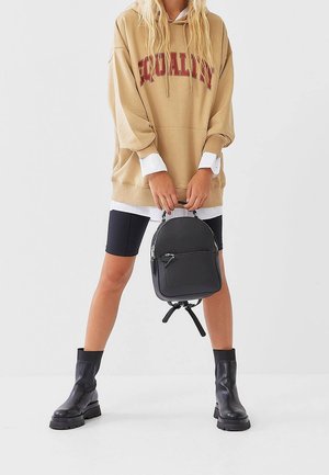 Osoba v béžové mikině s nápisem „EQUALITY“, v černých cyklistických šortkách a černých botách, držící malý černý batoh za horní rukojeť.