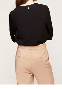 Svart långärmad topp med rund halsringning, som har en liten metalldetalj bak, i kombination med ljusbeiga skräddarsydda byxor.