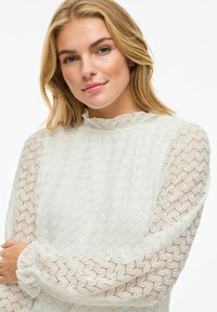 Blouse en dentelle blanche à motif texturé, avec un col haut et de longues manches légèrement bouffantes avec des poignets élastiques. Détails délicats sur l'ensemble.