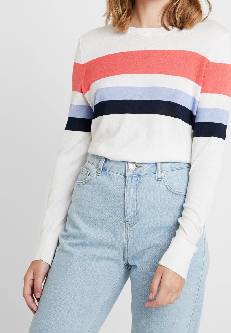 Femme portant un pull blanc à manches longues avec des rayures horizontales rouges, bleues et navy, rentré dans un jean taille haute bleu clair.