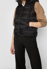 Gilet imbottito nero con zip frontale, cappuccio e sezioni trapuntate orizzontali. Presenta tasche laterali ed è indossato su un maglione beige e pantaloni neri.