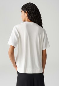 Haut blanc à manches courtes avec une texture lisse et des accents côtelés sur les manches. Ourlet droit et coupe décontractée, présenté de dos.