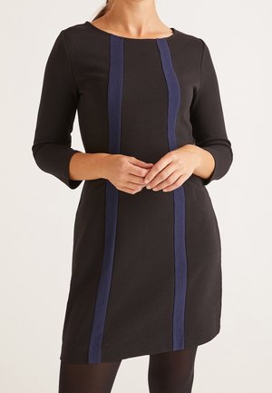 Mujer con un vestido negro hasta la rodilla, de manga tres cuartos y dos franjas verticales azul marino en el frente.