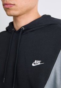 Young person wearing a black and gray Nike hoodie with the brand’s logo embroidered on the chest, visible from chin to upper torso.