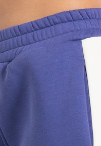 Pantalones deportivos morados con cintura elástica, textura de tela suave y una franja blanca decorativa a lo largo de la costura lateral.