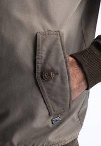 Béžová bunda s knoflíkovou náprsní kapsou, žebrovanými manžetami a textilní štítkem s logem "Petrol". Hladká, matná textura.