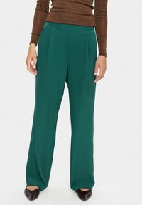 Pantalons larges verts en tissu lisse avec des plis à l'avant et une taille haute, associés à un haut à manches longues marron et des chaussures noires.