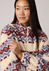 Φλις πουλόβερ σε κρεμ χρώμα με ζιγκ-ζαγκ μοτίβα σε μπορντό και μπλε. Διαθέτει ψηλό γιακά και κλείσιμο με φερμουάρ. Μαλακό, υφασμάτινο ύφασμα με υφή.