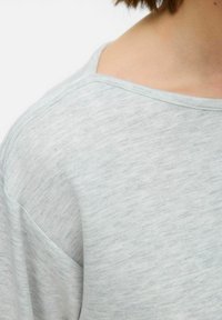 Gaiši pelēks, īsām piedurknēm tops ar plašu izgriezumu. Tam ir smalka svītraina struktūra un brīva piegriezuma siluets. Materiāls izskatās mīksts un ērts.