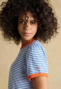 Frau mit lockigem Haar trägt ein blau-weiß gestreiftes T-Shirt mit orangem Saum und dem Schriftzug "JOULES" auf dem Ärmel, vor beigem Hintergrund.