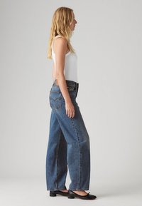 Pikendav blondi juustega naine, kellel on seljas valge ilma varrukateta topp, kõrge vöökoht ja laiad sinised teksad ning mustad madala kontsaga kingad, seismas profiilis.