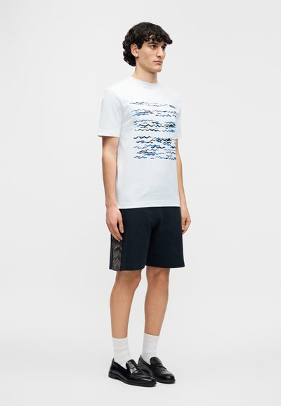 Λευκό βαμβακερό t-shirt με μπλε γραφικό κύμα, σε συνδυασμό με σκούρα σορτς που έχουν πλαϊνές πάνελ με σχέδιο. Το μοντέλο φοράει μαύρα loafers και λευκές κάλτσες.