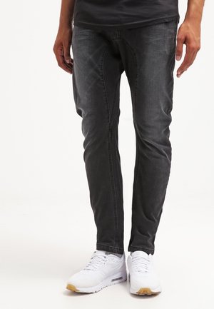 Person iført slim-fit sorte jeans og hvide sneakers, stående mod en ensartet lys baggrund.